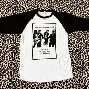 Puede incluir: Una camiseta de béisbol blanca con mangas negras de tres cuartos que presenta un estampado gráfico en blanco y negro de la banda The Heartbreakers. El texto dice THE HEARTBREAKERS y C.B.G.B.'s Fri., Sat., Sun. en un marco negro.