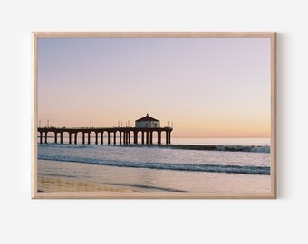 May include: A wooden framed photograph of a pier extending into the ocean at sunset. The pier has a small building at the end and people are walking on it. The sky is a soft pink and purple color.