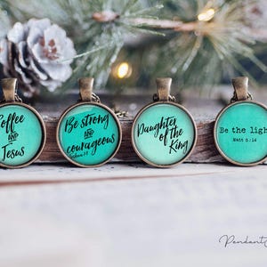 Puede incluir: Cuatro colgantes redondos de tono bronce con fondos verde azulado y texto en negro. Los colgantes dicen: Coffee and Jesus, Be strong and courageous Joshua 1:9, Daughter of the King y Be the light. Matt 5:14. Cada colgante tiene una pequeña anilla metálica.