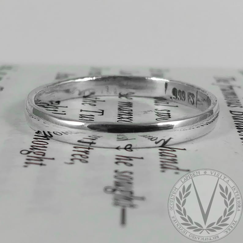 Puede incluir: Un anillo de plata con una superficie lisa y pulida. El anillo tiene un dise&ntilde;o sencillo y est&aacute; marcado con "999 FS". El anillo est&aacute; sobre un fondo de texto impreso.