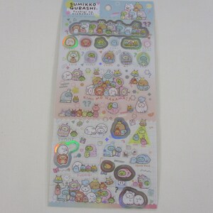 May include: A sheet of colorful, holographic stickers featuring characters from the Sumikko Gurashi franchise. The stickers feature a variety of characters, including a penguin, a bear, a cat, and a frog. The stickers are arranged in a grid pattern and are surrounded by a white border. The text "Sumikko Gurashi" and "Fushigi na o-tamago-dachi" is printed on the sheet.