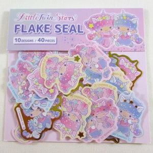 May include: A package of Little Twin Stars flake seals with 10 designs and 40 pieces. The seals feature the characters Kiki and Lala in various poses with a unicorn and stars. The package is pink and purple with gold accents.