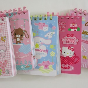 May include: A collection of spiral-bound notebooks with colorful covers featuring cartoon characters. The notebooks have pink, blue, and white covers with images of Hello Kitty, My Melody, and other cute characters. The spiral binding is pink or green.