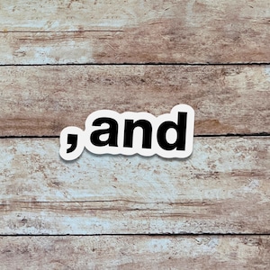 May include: A white sticker with the word ", and" in black, centered on a wooden background. The sticker has a thin white border. The wooden surface has a light brown color with visible wood grain.