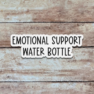 May include: A white sticker with the text "EMOTIONAL SUPPORT WATER BOTTLE" in a bold, black font. The sticker is placed on a wooden surface with a light brown color and visible wood grain.