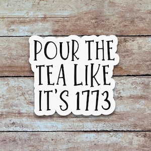 May include: A white sticker with black text that reads "POUR THE TEA LIKE IT'S 1773". The sticker has a rounded rectangular shape and is set against a wooden background.