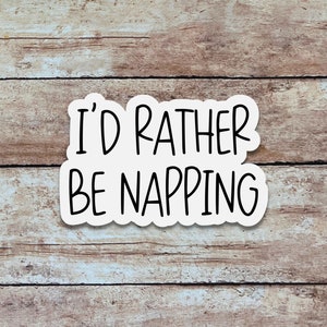 May include: A white sticker with the text "I'D RATHER BE NAPPING" in black, set against a wooden background. The sticker has a simple, rounded shape. The text is in a bold, easy-to-read font.