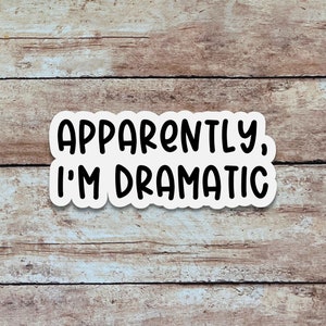 May include: A white sticker with the text "APPARENTLY, I'M DRAMATIC" in black, bold, uppercase letters. The sticker has a white border and is set against a wooden background with horizontal planks.
