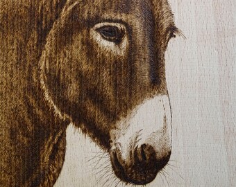 May include: A detailed wood-burned portrait of a donkey, showcasing the animal's profile. The artwork features a light brown background and a dark brown donkey with a white muzzle. The donkey's eye is visible, and the ears are erect.