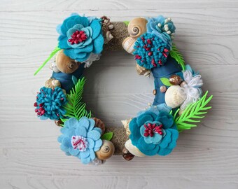 May include: A handcrafted wreath featuring a nautical theme. The wreath is adorned with turquoise felt flowers, seashells, and decorative elements. The design incorporates green fern-like accents and a sandy base, creating a coastal aesthetic.