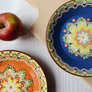 Può includere: Due piatti in ceramica decorativi con intricati disegni floreali. Il piatto più grande è blu con un fiore giallo e verde, mentre il piatto più piccolo è arancione con un fiore giallo e verde.