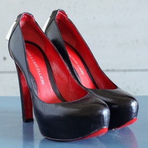 Op de afbeelding: Een paar zwarte pumps met hoge hakken en rode voering. De schoenen hebben een plateauzool en een spitse neus. De merknaam "Andrea Cancellieri" staat gedrukt aan de binnenkant van de ene schoen.