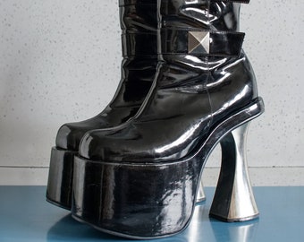 Könnte beinhalten: Ein Paar schwarze Lackplattformstiefel mit silbernen Schnallen und dicken Absätzen. Die Stiefel haben eine hohe Plattform und einen dicken Absatz.