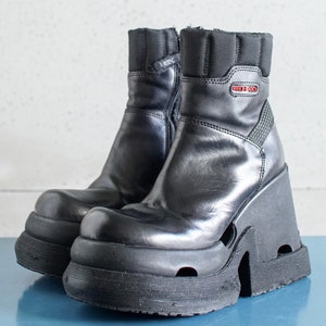 Può includere: Stivali platform in pelle nera con cerniera laterale e dettaglio a taglio sulla piattaforma. Gli stivali hanno un tacco spesso e massiccio e una suola testurizzata. Il nome del marchio "HEX" è visibile sul lato dello stivale.