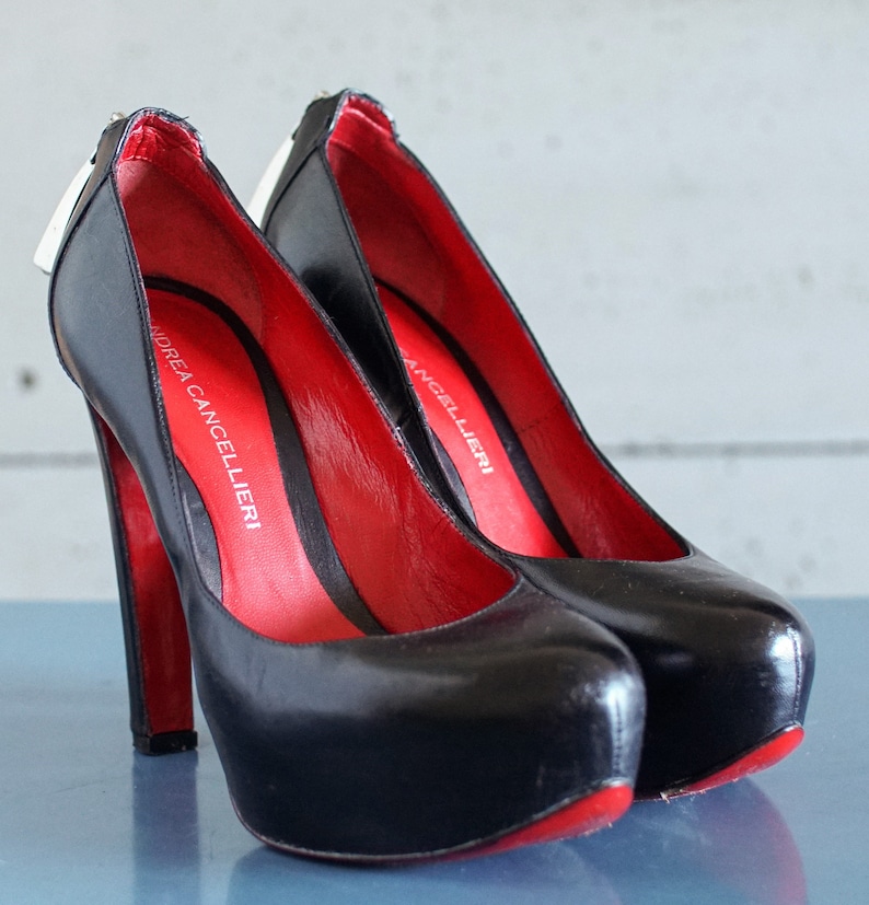 Op de afbeelding: Een paar zwarte pumps met hoge hakken en rode voering. De schoenen hebben een plateauzool en een spitse neus. De merknaam "Andrea Cancellieri" staat gedrukt aan de binnenkant van de ene schoen.