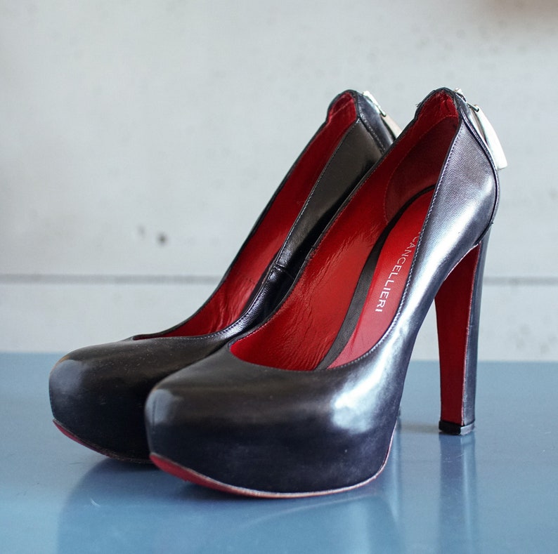 Op de afbeelding: Een paar zwarte leren pumps met hoge hakken en een rode voering. De schoenen hebben een rits aan de achterkant en de merknaam "CANCELLIERI" staat gedrukt aan de binnenkant van de schoen.