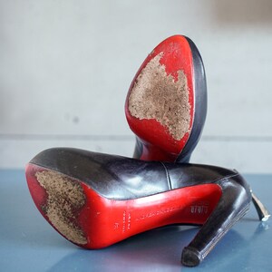 Op de afbeelding: Een paar zwarte leren hakken met rode zolen. De schoenen hebben een rits aan de zijkant en zijn licht beschadigd.
