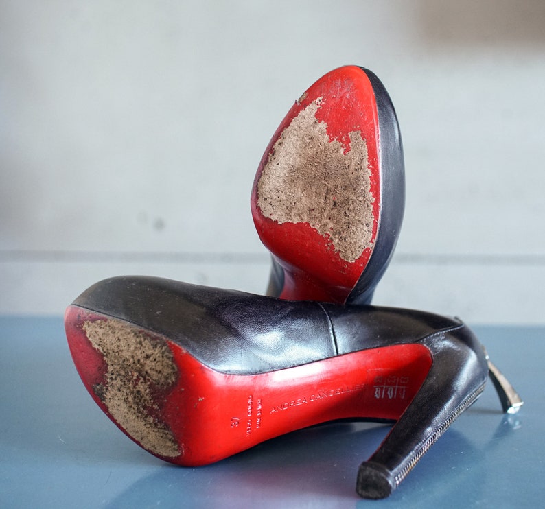 Op de afbeelding: Een paar zwarte leren hakken met rode zolen. De schoenen hebben een rits aan de zijkant en zijn licht beschadigd.