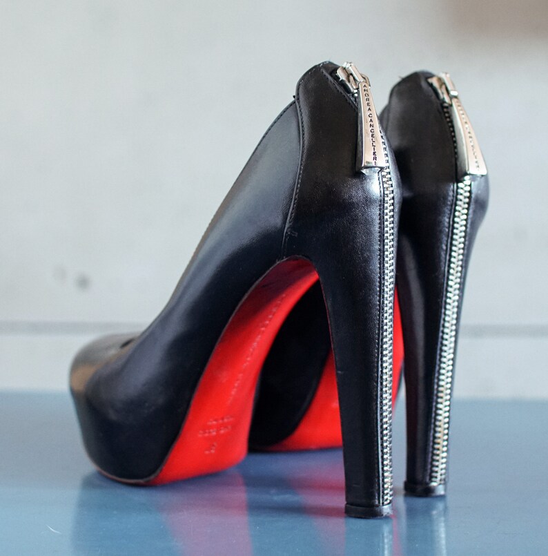 Op de afbeelding: Een paar zwarte leren pumps met hoge hakken, rode zolen en een ritsdetail aan de achterkant van elke schoen. De ritsen zijn zilverkleurig en hebben de tekst "SOFIA CANDELIER" erop.