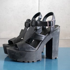 Könnte beinhalten: Ein Paar schwarze Plateausandalen mit dicken Blockabsätzen. Die Sandalen haben einen Knöchelriemen mit silberner Schnalle und eine strukturierte Sohle. Die Plateauhöhe beträgt ca. 5 cm.