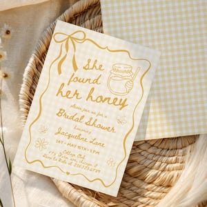 Op de afbeelding: Een uitnodiging voor een bruidsdouche met de tekst "She found her honey" in gouden letters. De uitnodiging staat op een crèmekleurige achtergrond met een gouden rand en een ruitpatroon. De uitnodiging ligt op een geweven dienblad met crèmekleurige stof.