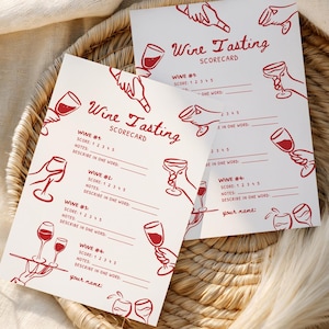 Puede incluir: Dos tarjetas blancas de cata de vinos con ilustraciones y texto en rojo, incluyendo las palabras "Wine Tasting SCORECARD". Las tarjetas están sobre una bandeja tejida con un fondo de tela blanca. Las tarjetas tienen espacio para notas.