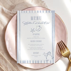May include: A menu card with a light blue and white striped border sits on a pink plate. The menu reads "Sailor's Beach Club" with starter, main course, and dessert options. A gold fork and knife are crossed on the plate.