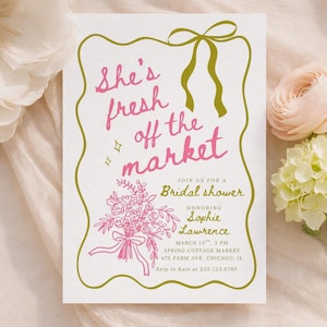 以下が含まれることがあります： ピンク色の筆記体で「She's fresh off the market」と書かれたブライダルシャワーの招待状。緑色の縁とリボンで縁取られています。招待状には花のイラストとイベントの詳細が含まれています。