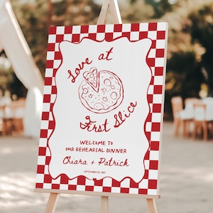 Op de afbeelding: Een rechthoekig bord op een houten ezel met een rood-wit geblokte rand. Het midden toont een rode lijntekening van een pizza met de tekst Love at First Slice, Welcome to our Rehearsal Dinner, Chiara + Patrick, 15 september 2023.