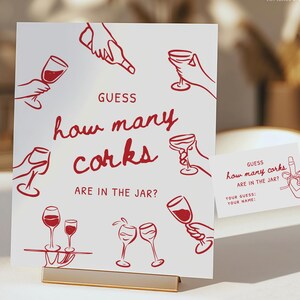 May include: A white card with red line drawings of wine glasses and hands holding wine bottles. The text "GUESS how many corks ARE IN THE JAR?" is written in red. A smaller card with the same design is also present.