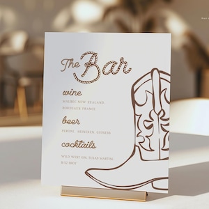 Puede incluir: Una tarjeta blanca con el texto "The Bar" en una fuente decorativa, con una ilustración de una bota de vaquero. La tarjeta enumera opciones de vino, cerveza y cócteles, incluyendo Malbec, Peroni y Texas Martini.