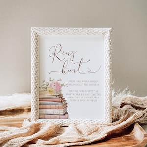 May include: A white frame with a white background and brown text that reads "Ring Hunt". The text is surrounded by a floral illustration and a stack of books. The text below the title reads "There are rings hidden throughout the shower. The one who finds the most rings by the time the first gift is unwrapped wins a special prize."