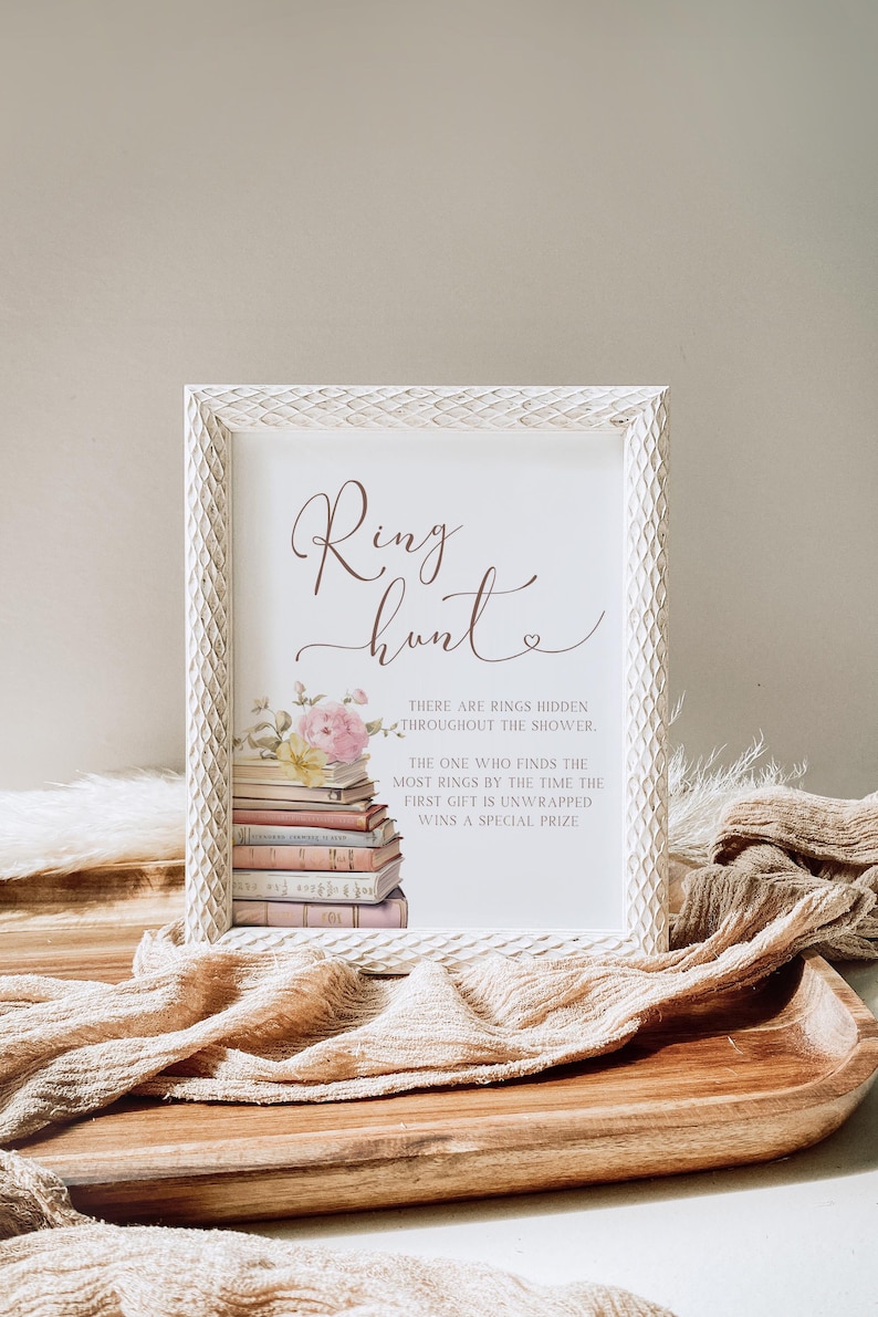 May include: A white frame with a white background and brown text that reads "Ring Hunt". The text is surrounded by a floral illustration and a stack of books. The text below the title reads "There are rings hidden throughout the shower. The one who finds the most rings by the time the first gift is unwrapped wins a special prize."