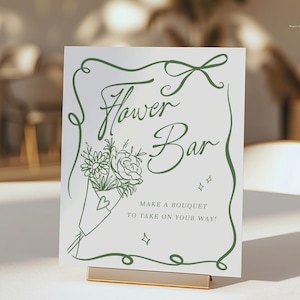 Puede incluir: Un letrero blanco con texto e ilustraciones verdes para una barra de flores. El letrero dice "Flower Bar" con una ilustración de ramo y el texto "Make a bouquet to take on your way!". El letrero está sujeto por un soporte de metal dorado.