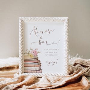 May include: White sign with a brown script font that says "Mimosa Bar" with a small heart. The sign has a watercolor illustration of a stack of books with pink flowers on top. The text below the illustration says "Pour the bubbly, add some juice, top with fruit, enjoy!"