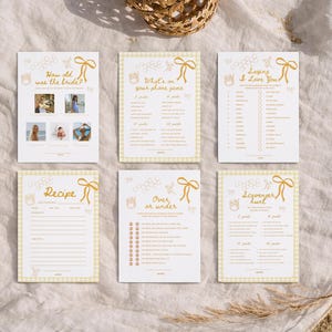 K&ouml;nnte beinhalten: Sechs wei&szlig;e Karten mit goldenem Text und einem gelben Vichy-Rand sind auf einem wei&szlig;en Tuch angeordnet. Die Karten tragen Titel wie "How old was the bride?" und "Recipe". Auf jeder Karte befindet sich eine dekorative Schleife und eine Bienen-Grafik.
