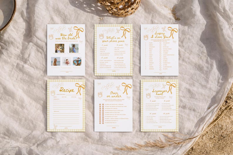 K&ouml;nnte beinhalten: Sechs wei&szlig;e Karten mit goldenem Text und einem gelben Vichy-Rand sind auf einem wei&szlig;en Tuch angeordnet. Die Karten tragen Titel wie "How old was the bride?" und "Recipe". Auf jeder Karte befindet sich eine dekorative Schleife und eine Bienen-Grafik.