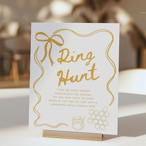 K&ouml;nnte beinhalten: Ein wei&szlig;es Schild mit goldfarbener Aufschrift "Ring Hunt". Das Schild hat eine goldene Schleife und einen dekorativen Rand. Unter dem Titel steht Text &uuml;ber ein Ring-Suchspiel. Ein goldener St&auml;nder h&auml;lt das Schild.