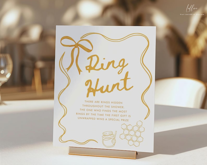 K&ouml;nnte beinhalten: Ein wei&szlig;es Schild mit goldfarbener Aufschrift "Ring Hunt". Das Schild hat eine goldene Schleife und einen dekorativen Rand. Unter dem Titel steht Text &uuml;ber ein Ring-Suchspiel. Ein goldener St&auml;nder h&auml;lt das Schild.
