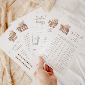 May include: A set of bridal shower game cards with a book stack and floral design. The cards include "Would She Rather", "Bridal Bingo", and "Never Have I Ever".