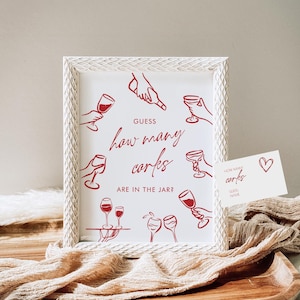 May include: White framed sign with red text and illustrations. The sign reads "Guess how many corks are in the jar?" and features drawings of hands holding wine glasses. A small card with the same text and a heart is also present.