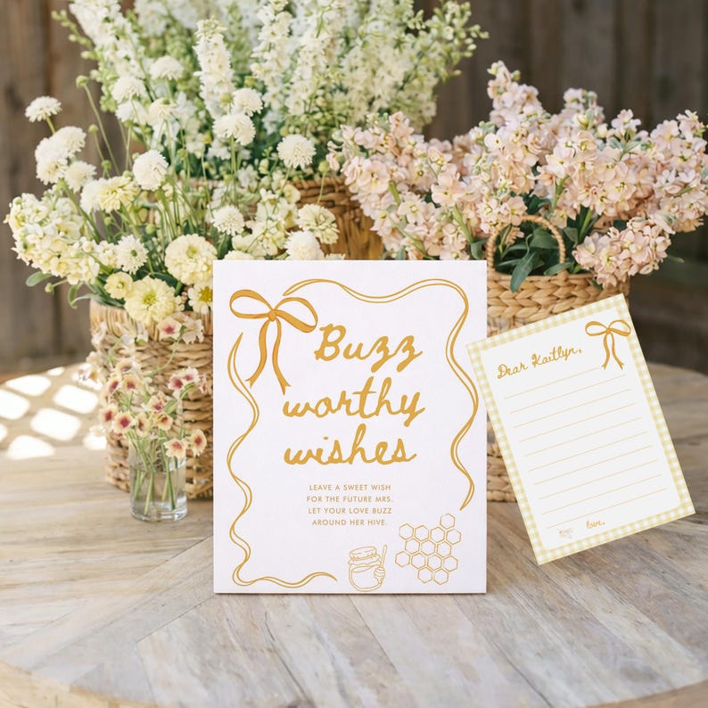 K&ouml;nnte beinhalten: Ein wei&szlig;es Schild mit goldenem Text, der "Buzz worthy wishes" mit einem Schleifendesign zeigt. Eine kleinere Karte mit dem Text "Dear Kaitlyn" befindet sich neben dem Schild. Blumenk&ouml;rbe sind im Hintergrund.