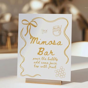 K&ouml;nnte beinhalten: Ein wei&szlig;es Schild mit goldfarbenen Buchstaben und Illustrationen f&uuml;r eine Mimosa-Bar. Das Schild zeigt eine Schleife, eine Biene, ein Honigglas und ein Wabenmuster. Der Text lautet "Mimosa Bar" und enth&auml;lt Anweisungen.