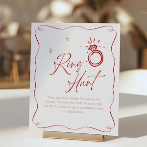 May include: A white sign with red text and a decorative border. The sign reads "Ring Hunt" with an illustration of a diamond ring. Text below explains the game rules. The sign is held by a gold-colored metal stand.