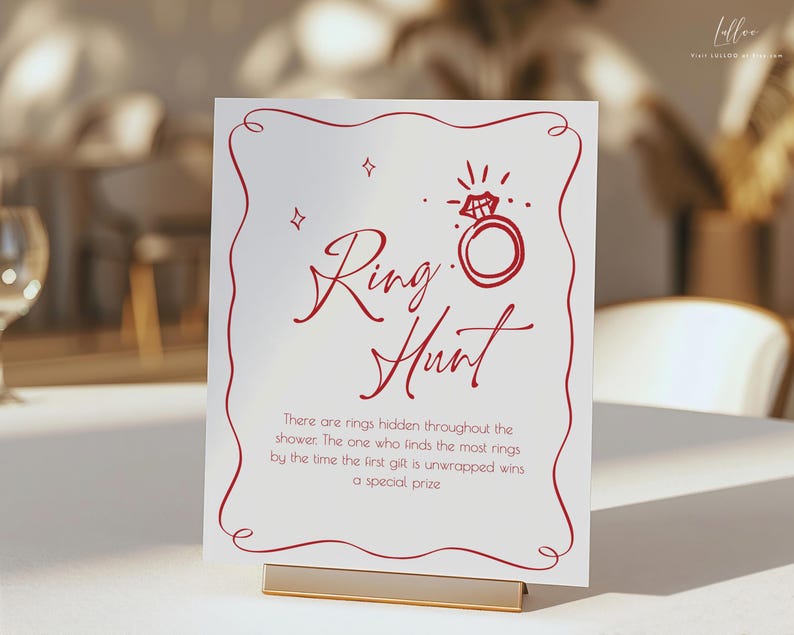 May include: A white sign with red text and a decorative border. The sign reads "Ring Hunt" with an illustration of a diamond ring. Text below explains the game rules. The sign is held by a gold-colored metal stand.