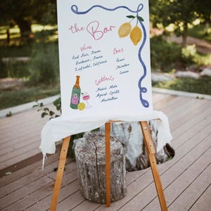 May include: A white sign with blue and pink lettering on an easel, listing the drinks available at the bar. The sign says "the Bar" and lists wine, beer, and cocktails. The sign is decorated with lemons and a bottle of champagne.