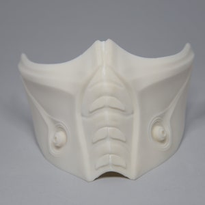Może przedstawiać: Biała plastikowa maska z detalicznym wzorem, przypominająca maskę na twarz z ostrym podbródkiem i wypukłym, fakturowanym grzbietem w środku.