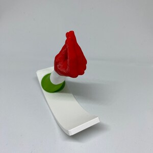 K&ouml;nnte beinhalten: Eine rote 3D-gedruckte Handskulptur in der italienischen Handgeste "mano fico" oder "figa". Die Hand ist auf einem wei&szlig;en und gr&uuml;nen Sockel montiert.