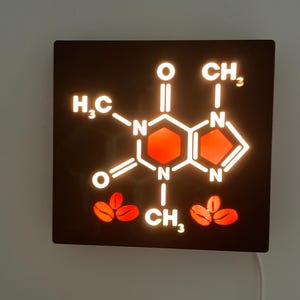 Könnte beinhalten: Ein quadratisches, schwarzes Wandkunstwerk mit einem leuchtenden weißen und roten Koffeinmolekül-Design. Die Molekularstruktur ist beleuchtet, mit Kaffeebohnen-Illustrationen am unteren Rand. Ein weißes Netzkabel ist sichtbar.