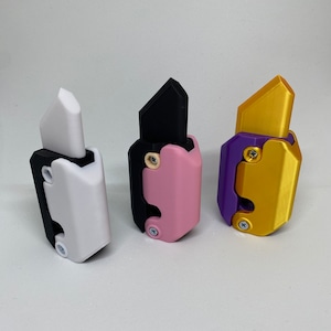 Könnte beinhalten: Drei kleine, handliche Werkzeuge mit einem einzigartigen Design. Die Werkzeuge sind aus Kunststoff und haben ein schwarzes, rosa und goldenes Farbschema. Jedes Werkzeug hat eine Klinge, die sich in den Griff einklappen lässt.